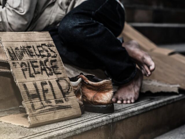 a homeless person of colour lies on some steps by a sign reading "homeless - please help"