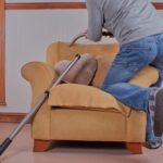 a man kneels on an armchair as he hoovers its upholstery