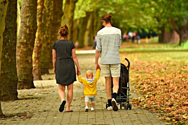 a mother and father walk with a child between them, holding their hands, in a park in autumn